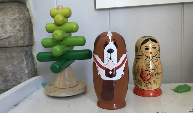 Three wooden toys sit on a white wooden shelf. In the center, a nesting doll painted to look like a St. Bernard dog. On the right, a faded nesting doll painted in a traditional style, painted as a smiling woman with a headscarf and floral dress. On the left, a third wooden stacking toy for toddlers. 