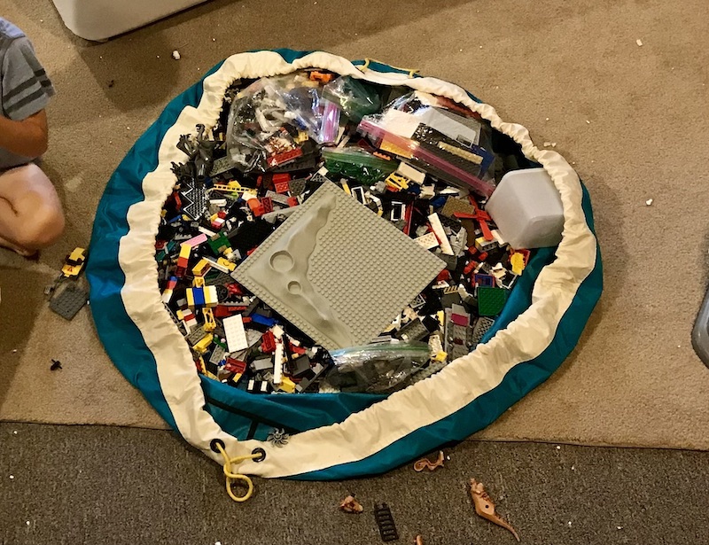 A nylon drawstring bag is opened up wide on a carpeted floor, displaying hundreds of Lego bricks and a gray Lego baseplate.