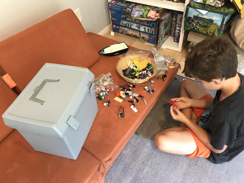 Small piles of Lego bricks are strewn along an orange cushion, where an 8-year-old boy sits inspecting a red Lego brick. A gray tackle box about one foot long and two feet wide sits next to the piles of Lego. 