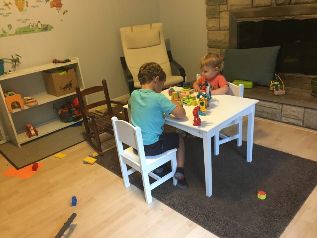Two little boys playing with toys at a while, child-sized table. A few feet away against a wall is a white shelving unit about three feet high. The shelf contains a basket of blocks and some colorful wooden toddler toys.